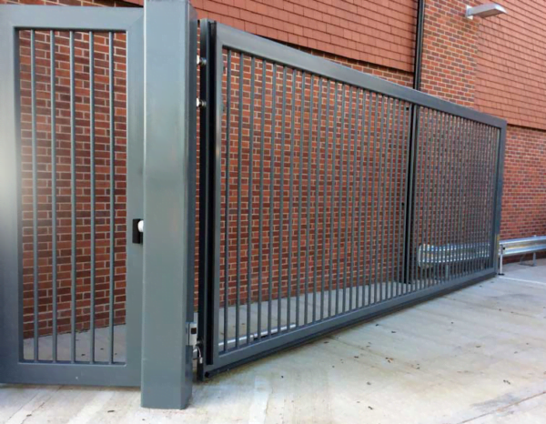 Velká šedá posuvná kovová brána s vertikálními příčkami a menší brankou u cihlové zdi.