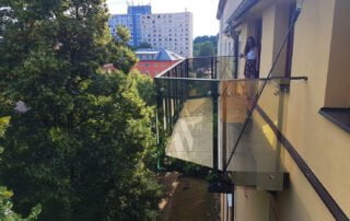 Skleněný balkon s kovovou konstrukcí Moderní skleněný balkon s ocelovou konstrukcí na žluté fasádě domu, realizace KOVKOM.