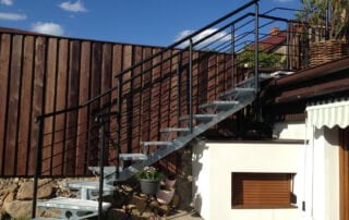 Ocelové schody se zábradlím Venkovní ocelové schodiště s pozinkovanými stupni a černým zábradlím vedoucí na terasu. Zakázková výroba KOVKOM.