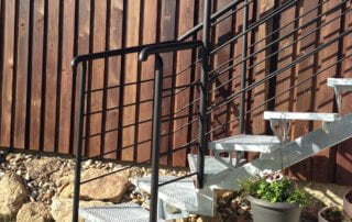 Realizace kovové schodiště Venkovní kovové schodiště s černým zábradlím a pozinkovanými roštovými stupni u dřevěného plotu.