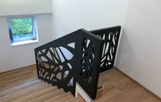 Zakázkové černé kovové zábradlí s moderním laserem řezaným vzorem pro interiérové schodiště. Realizace KOVKOM.