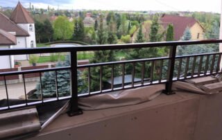 Kovové balkonové zábradlí instalace Nově instalované černé kovové zábradlí na balkoně s výhledem do zelené zahrady a okolní zástavby.