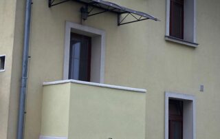 Zakázkový přístřešek s ocelovou konstrukcí Zakázkový přístřešek z polykarbonátu s černou ocelovou konstrukcí nad oknem a balkonem na fasádě domu.