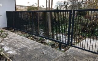 Moderní tmavě šedý kovový plot s vertikálními tyčemi a brankou, instalovaný u zámkové dlažby. Realizace KOVKOM.