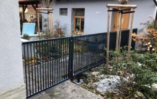 Kovový plot na zakázku Moderní černý kovový plot s vertikálními tyčemi, instalovaný u chodníku a zahrady rodinného domu.